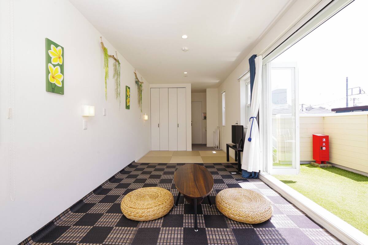 A hallway with a checkered floor, a wooden table, two woven stools, a door, and a view of a garden outside.