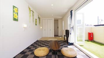 A hallway with a checkered floor, a wooden table, two woven stools, a door, and a view of a garden outside.