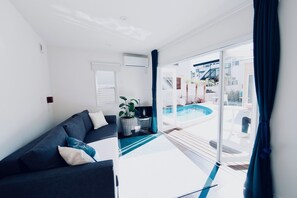 Un salón moderno con un sofá oscuro, una mesita y una planta en maceta. Un ventanal corredero da a un área exterior con piscina.