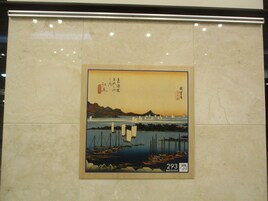 Uma xilogravura japonesa emoldurada com uma cena costeira, incluindo barcos e montanhas, exposta numa parede de mármore.
