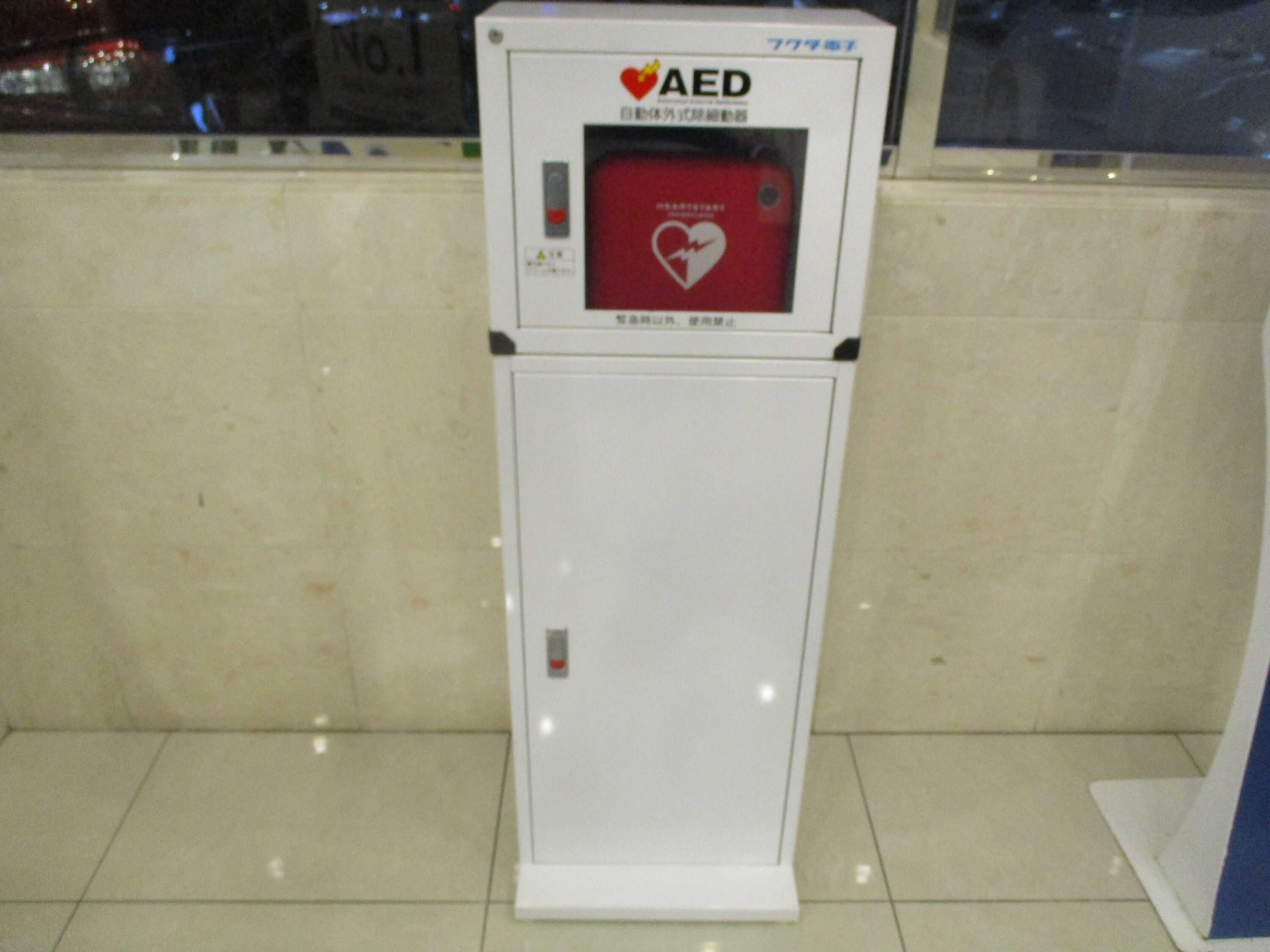 A white AED emergency medical device with a red panel and a heart symbol, placed on a tiled floor against a wall.
