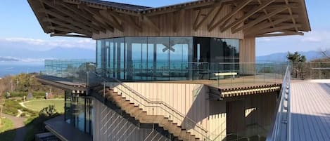 A modern building with a glass enclosure, wooden deck, and a staircase leading to an upper level.