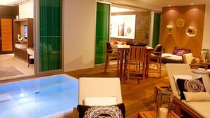 A modern hotel room with a glass-enclosed pool, a dining area with a table and chairs, and a seating area with sofas and armchairs.