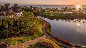 A resort with a curved pathway leading to a body of water, surrounded by lush greenery and palm trees.