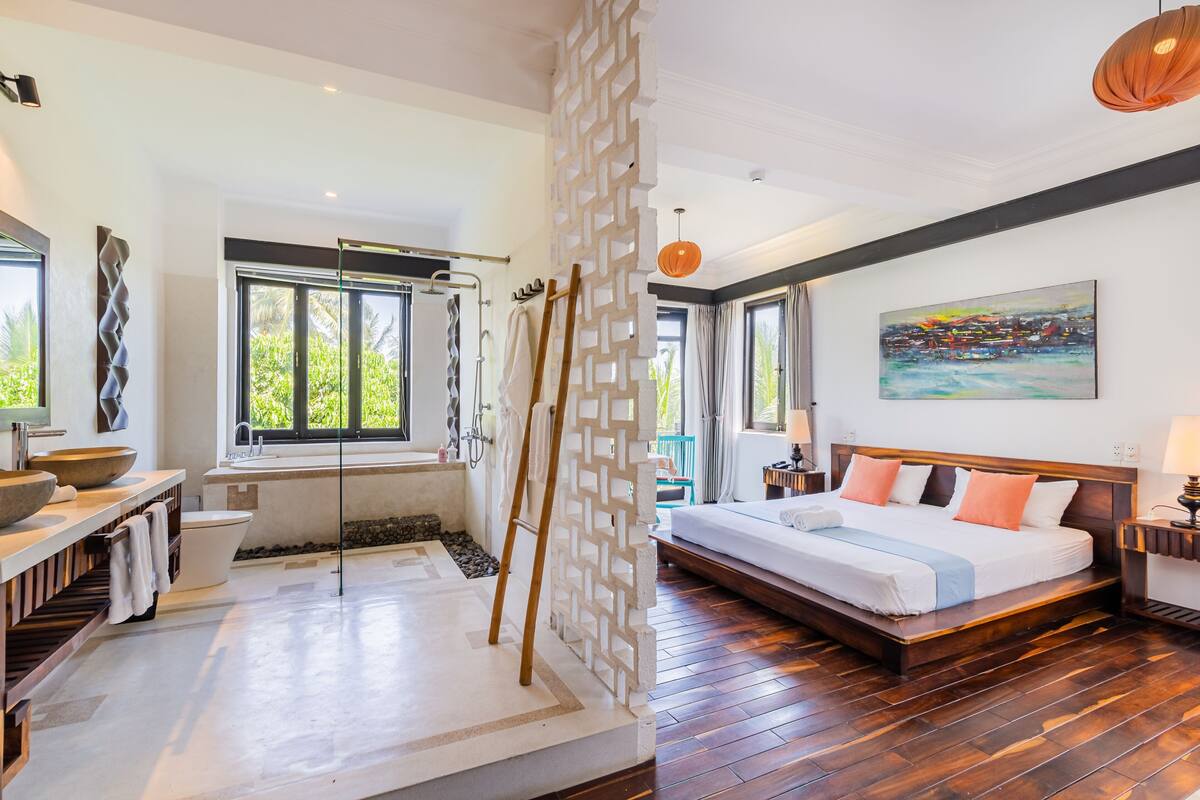 A modern hotel room with a large bed, a bathtub, a wooden floor, and a view of greenery outside.