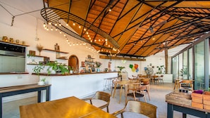 Restaurante com mesas e cadeiras de madeira, balcão com plantas e teto com vigas aparentes e luzes pendentes.