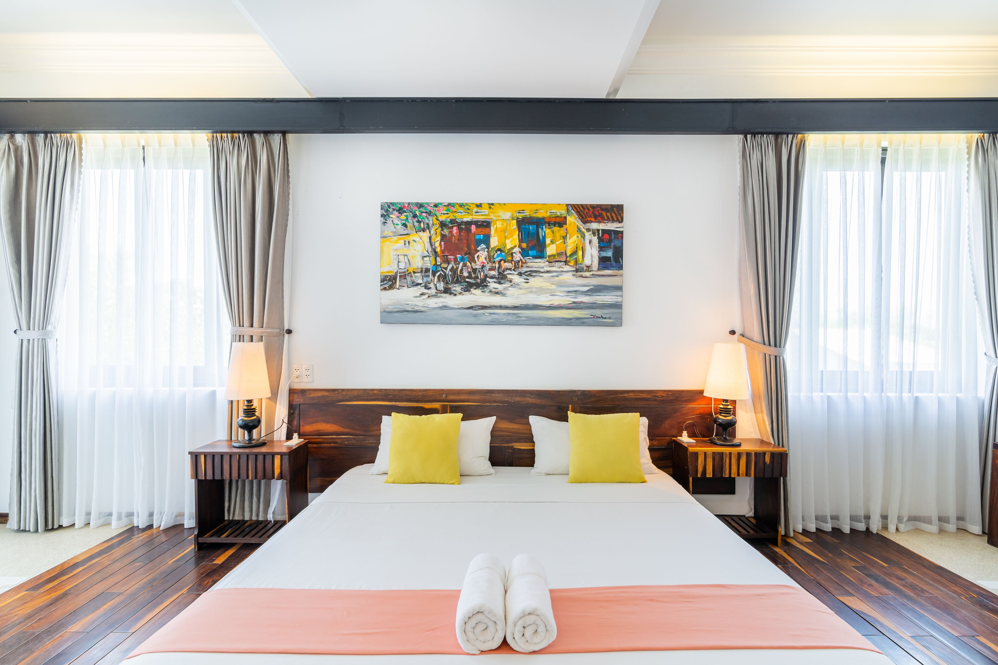 A hotel room with a large bed, two bedside tables with lamps, a painting on the wall, and curtains on the windows.