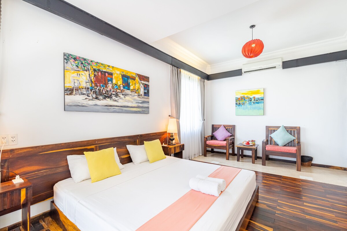 A hotel room with a large bed, wooden headboard, two chairs, a small table, and artwork on the walls.