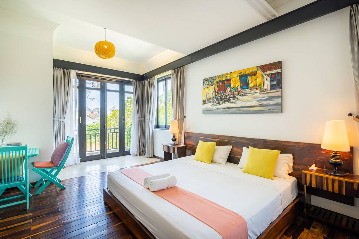 A hotel room with a large bed, a wooden headboard, a painting on the wall, and a view of the outdoors through the windows.