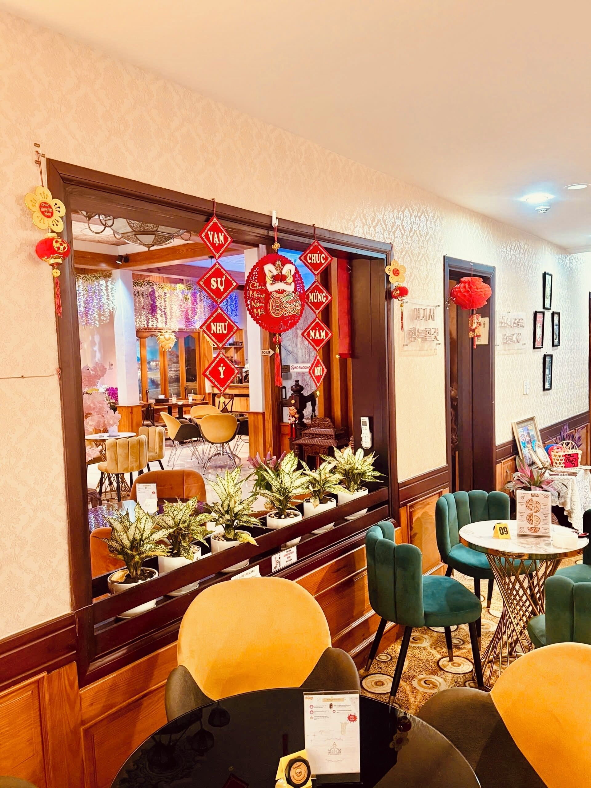 Un vestíbulo de hotel con un espejo grande que refleja el interior, decorado con adornos rojos de Año Nuevo chino. Hay plantas en macetas sobre una mesa, sillas tapizadas de verde y una pequeña mesa redonda con un menú.