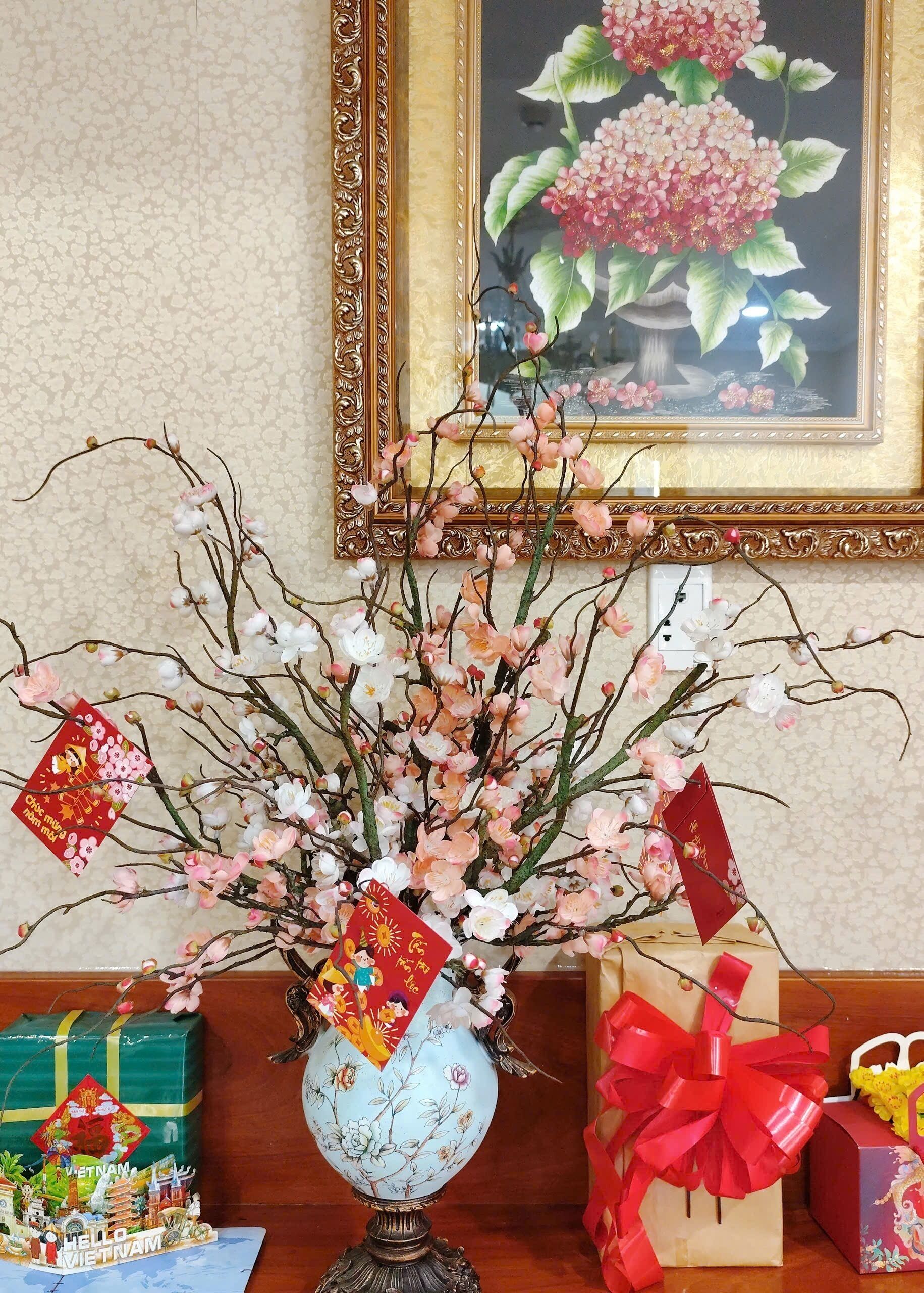 A decorative vase with artificial flowers and red tags, placed on a table with wrapped gifts and a framed painting in the background.