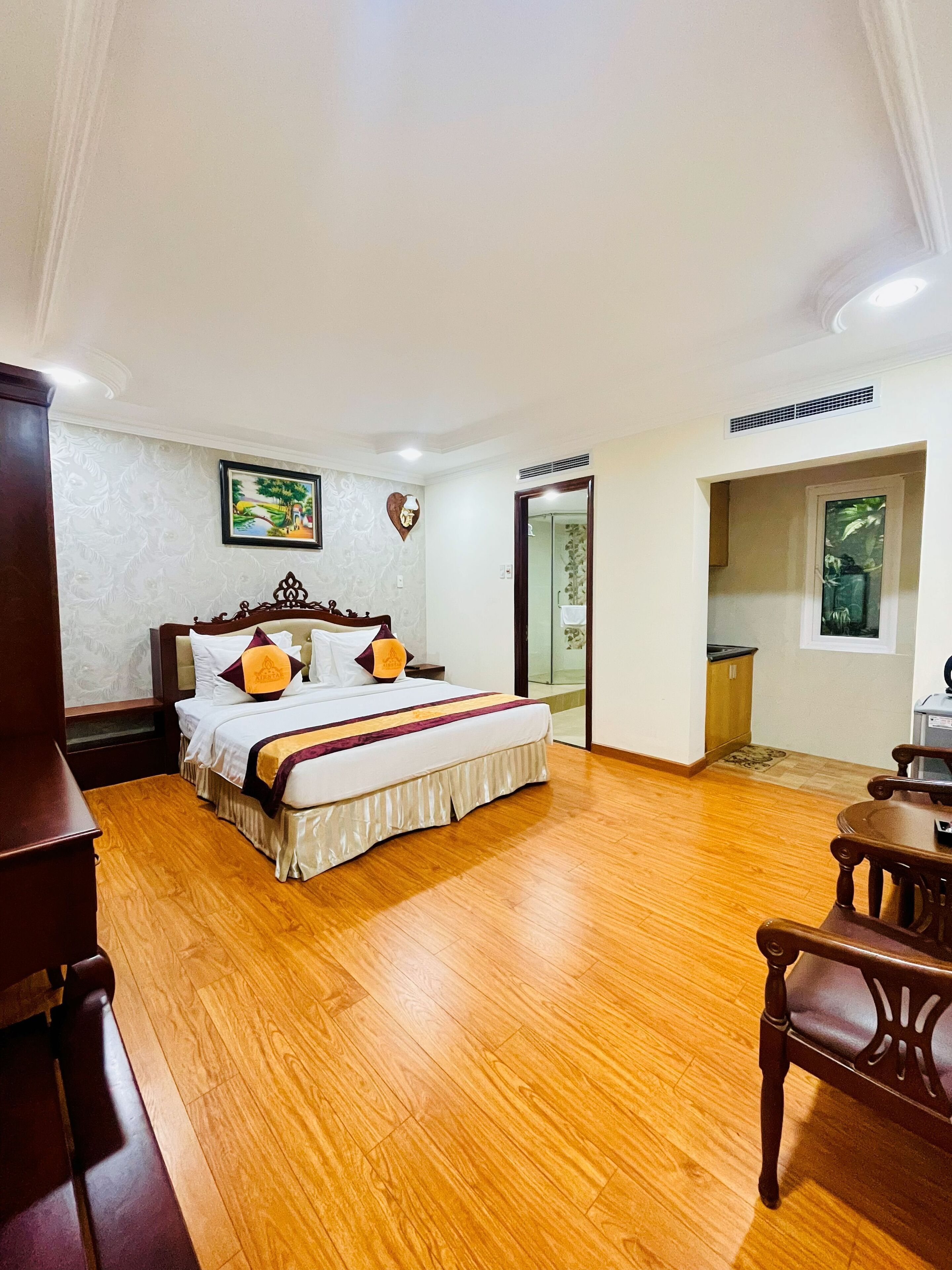 A hotel room with a large bed, wooden flooring, a wooden nightstand, a chair, a small table, and a window with a view of greenery.