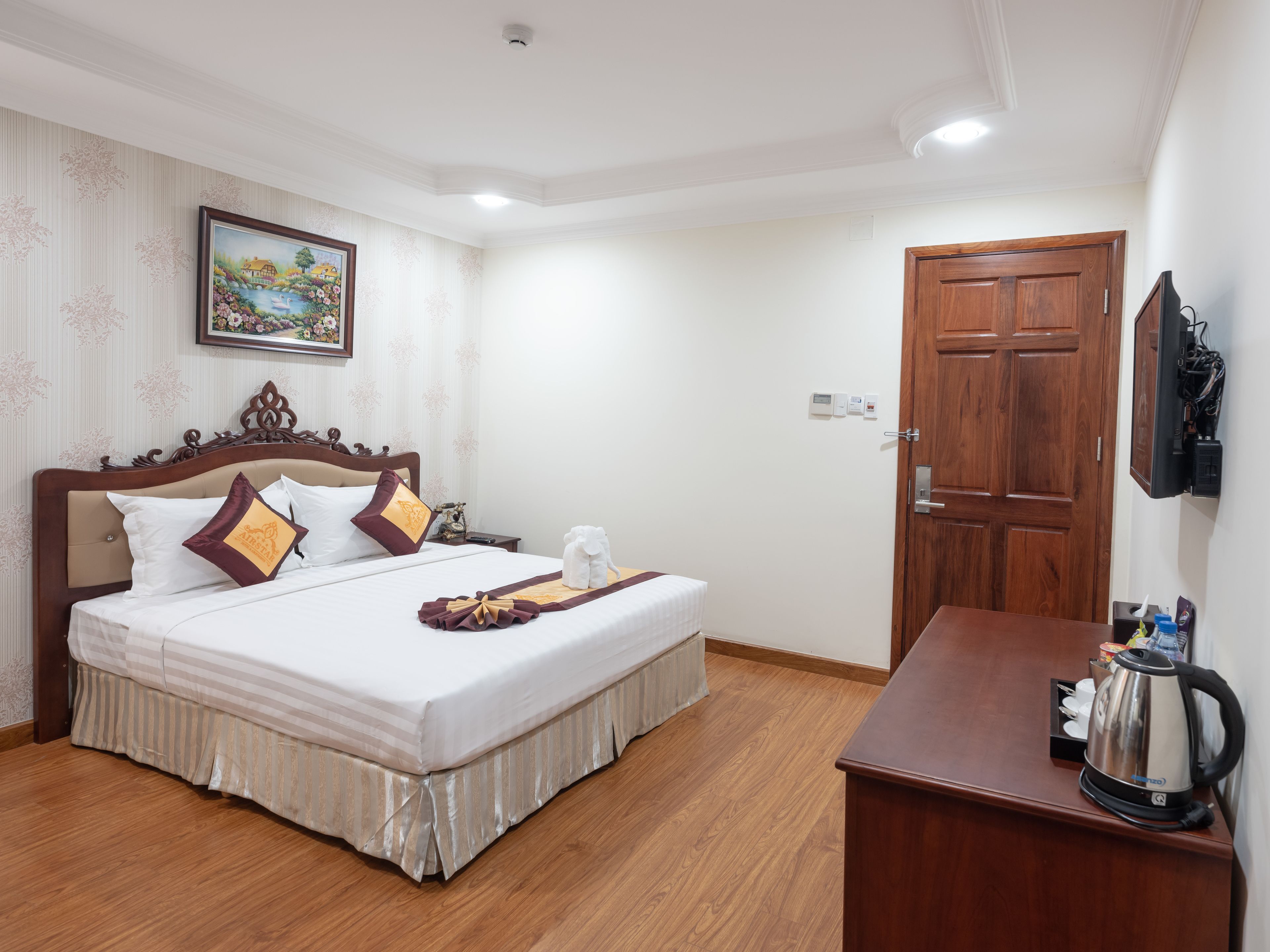 A hotel room with a large bed, a wooden headboard, a painting on the wall, a wooden door, and a small table with a kettle.
