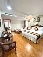 A hotel room with two single beds, a wooden chair, a small table with a bottle and glasses, and a painting on the wall.