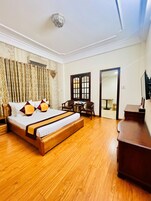 Quarto de hotel com cama de madeira, uma mesa e uma cadeira. Há um espelho na parede e uma porta que dá para outro quarto.