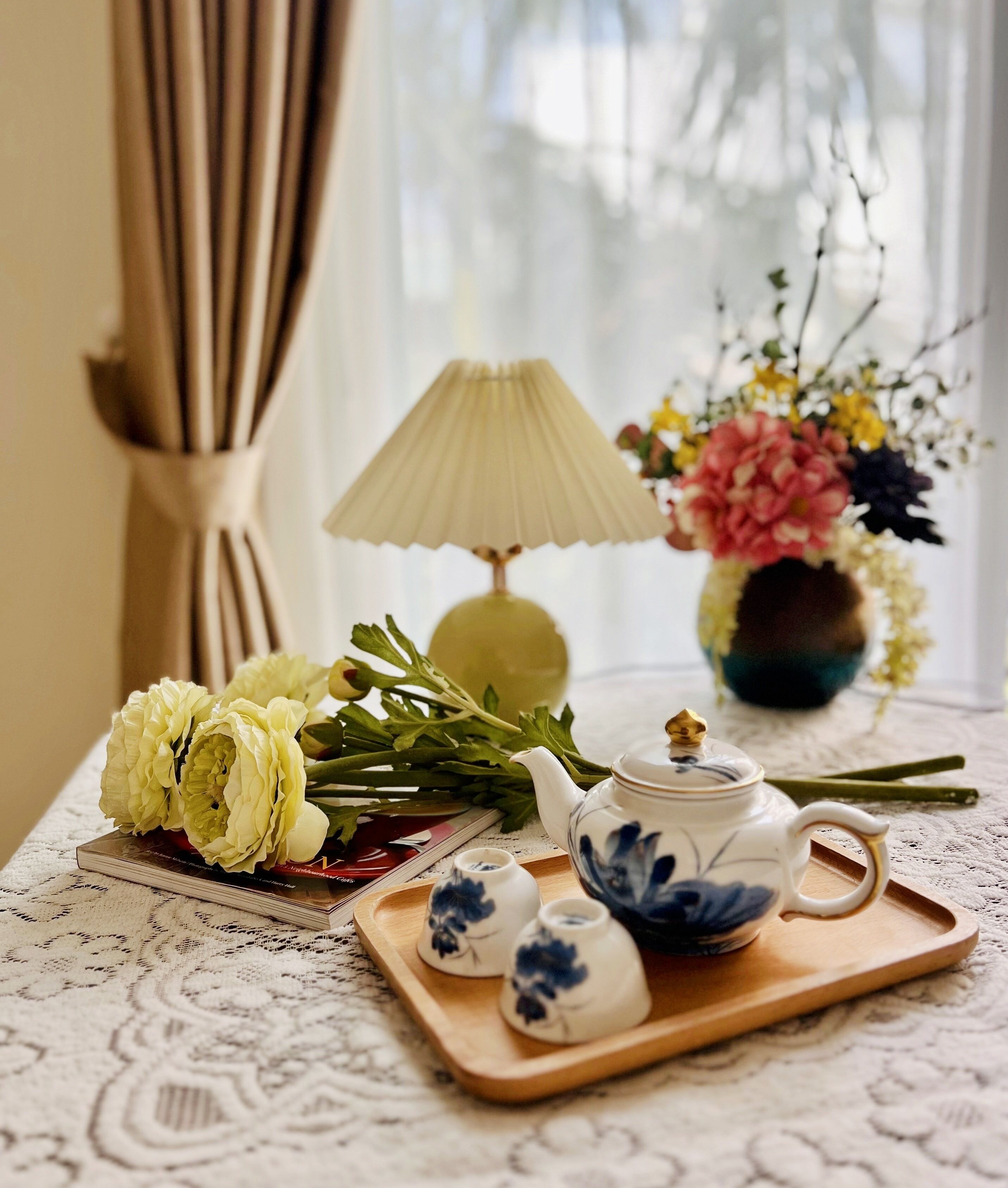Una mesa con una tetera, tazas y flores en una bandeja, una lámpara y un jarrón de flores.