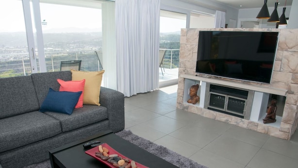 A modern living room with a grey sofa, colorful cushions, a black coffee table with decorative items, a large flat-screen TV, and a stone fireplace. - 6 Barquentine (Knysna)