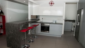 A modern kitchen with a central island, bar stools, a microwave, oven, and a washing machine. - 6 Barquentine (Knysna)