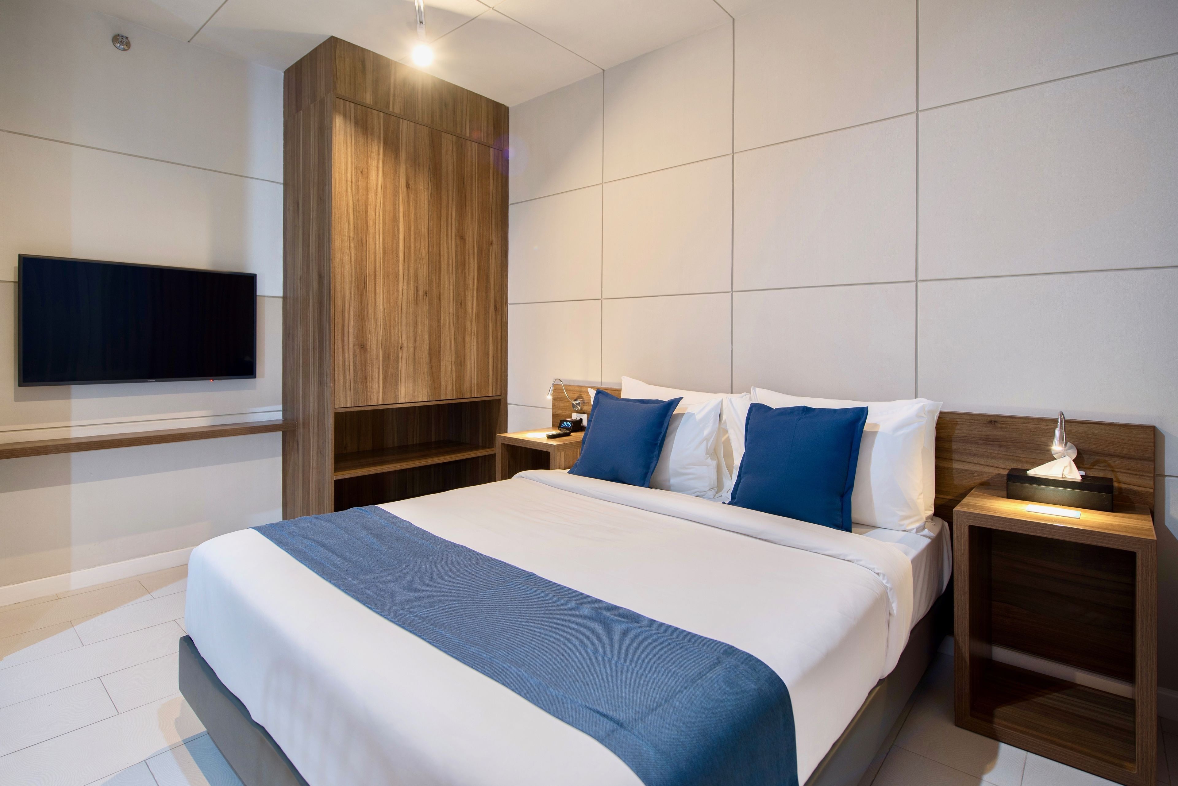 A modern hotel room with a large bed, a flat-screen TV mounted on the wall, wooden paneling, and a nightstand with a lamp.