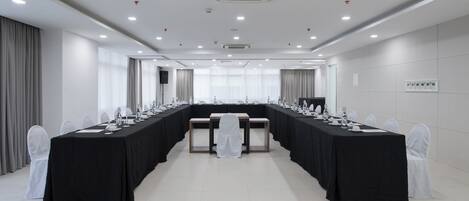 Un salón de conferencias con una larga mesa, sillas blancas y un mantel blanco.