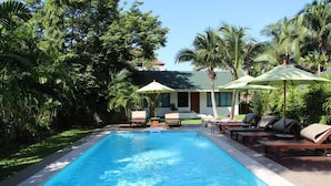 A swimming pool with lounge chairs and umbrellas, surrounded by palm trees and greenery. - Villa Raeya (Krabi)