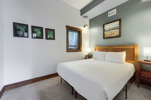 Ein Hotelzimmer mit einem großen Bett, einem Nachttisch, einer Lampe, einem Fenster mit Jalousien und gerahmten Bildern an der Wand.