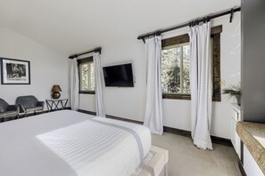 A bedroom with a large bed, two windows with curtains, a TV on the wall, a desk with a chair, and a plant.