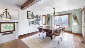 Un coin repas avec une grande table en bois, des chaises et un lustre. Les murs sont ornés de tableaux encadrés, et une fenêtre offre une vue sur les arbres.