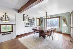 Ein Essbereich mit einem großen Holztisch, Stühlen und einer Chandelier. An der Wand befinden sich gerahmte Bilder und ein Fenster mit Blick auf Bäume.