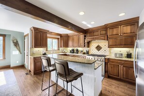 A kitchen with wooden cabinets, a granite countertop, a central island with bar stools, a stainless steel refrigerator, and a window providing natural light. - Free SkyCard Activi - Close To Town/Lifts, Private Hot Tub - High Point Chateau (Breckenridge)