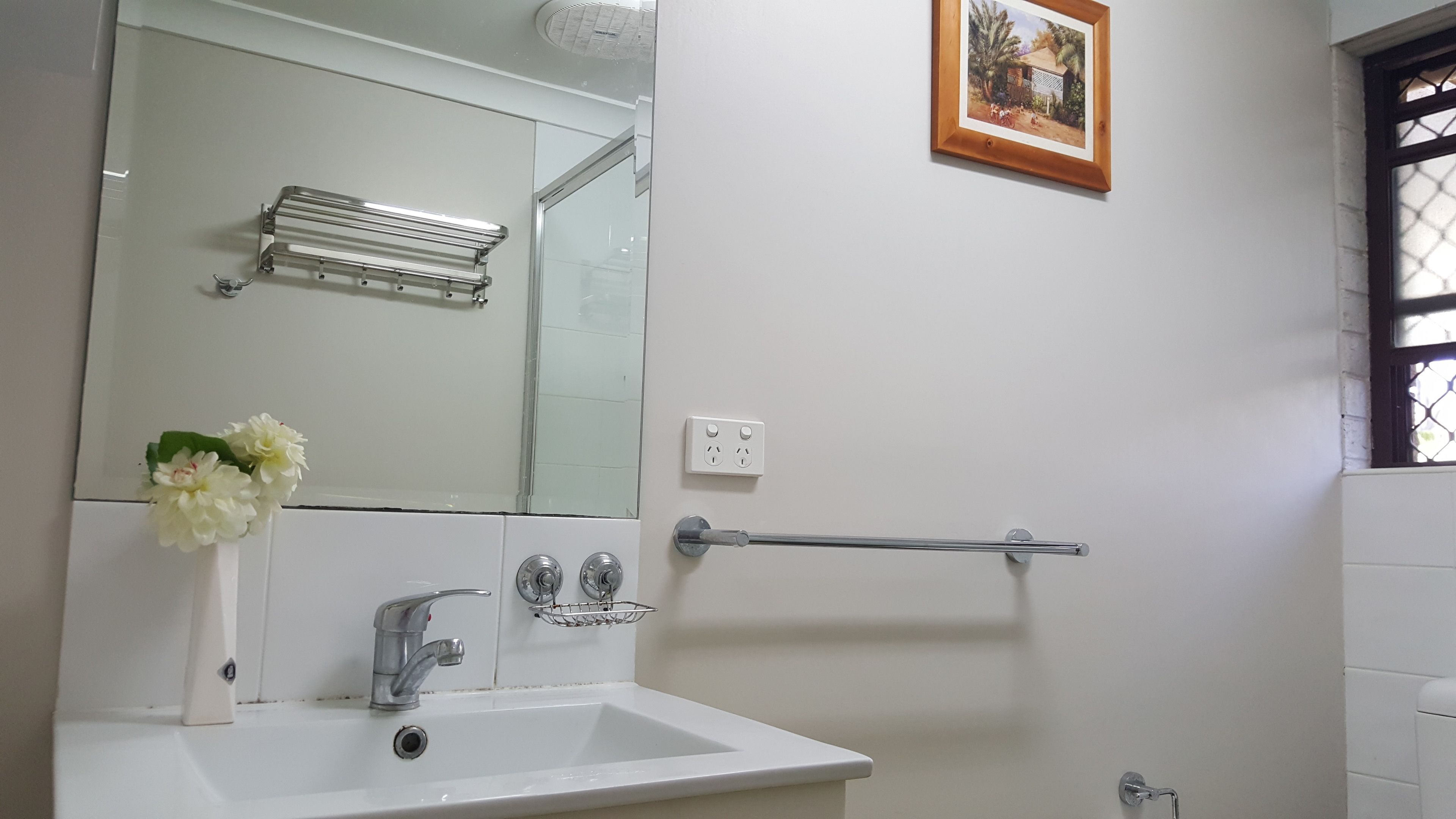 Un baño con lavamanos blanco, espejo y toallero. Hay una foto en la pared y una ventana con reja de metal.