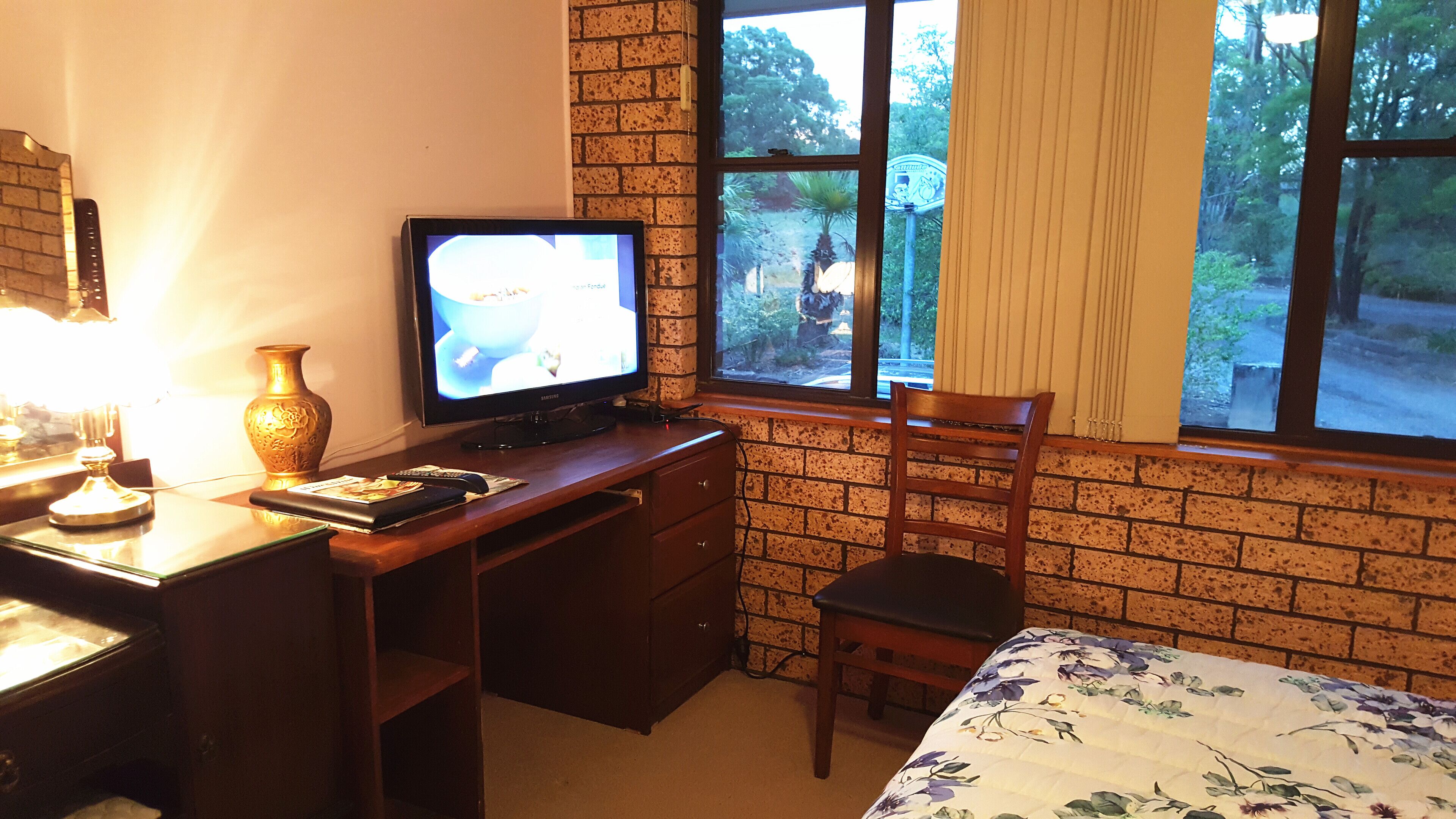 A room with a brick wall, a desk with a television, a chair, and a window with a view of trees.