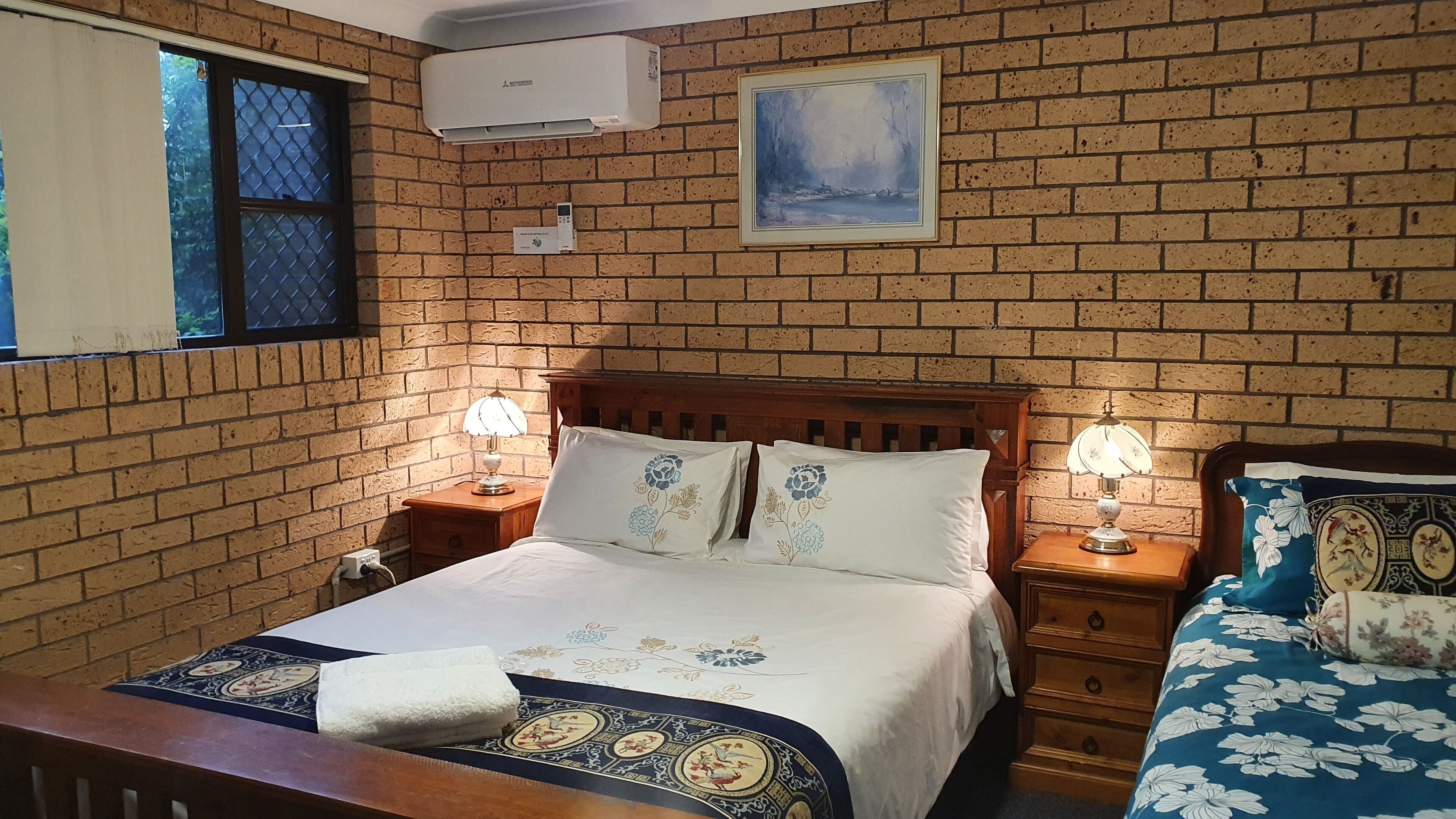 A bedroom with a brick wall, a bed with floral bedding, two bedside tables with lamps, a window with a view, and an air conditioning unit.
