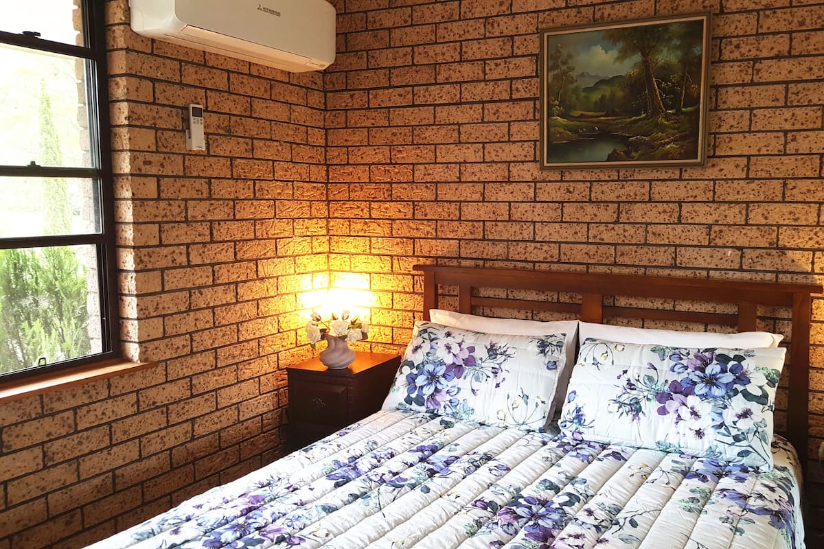 Ein Schlafzimmer mit Backsteinwand, einem Bild, einem Bett mit floralem Bettzeug, einem Nachttisch mit Lampe und einem Fenster mit Blick auf Bäume.