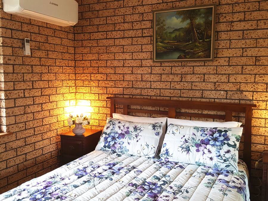 A bedroom with a brick wall, a painting, a bed with floral bedding, a nightstand with a lamp, and a window with a view of trees.