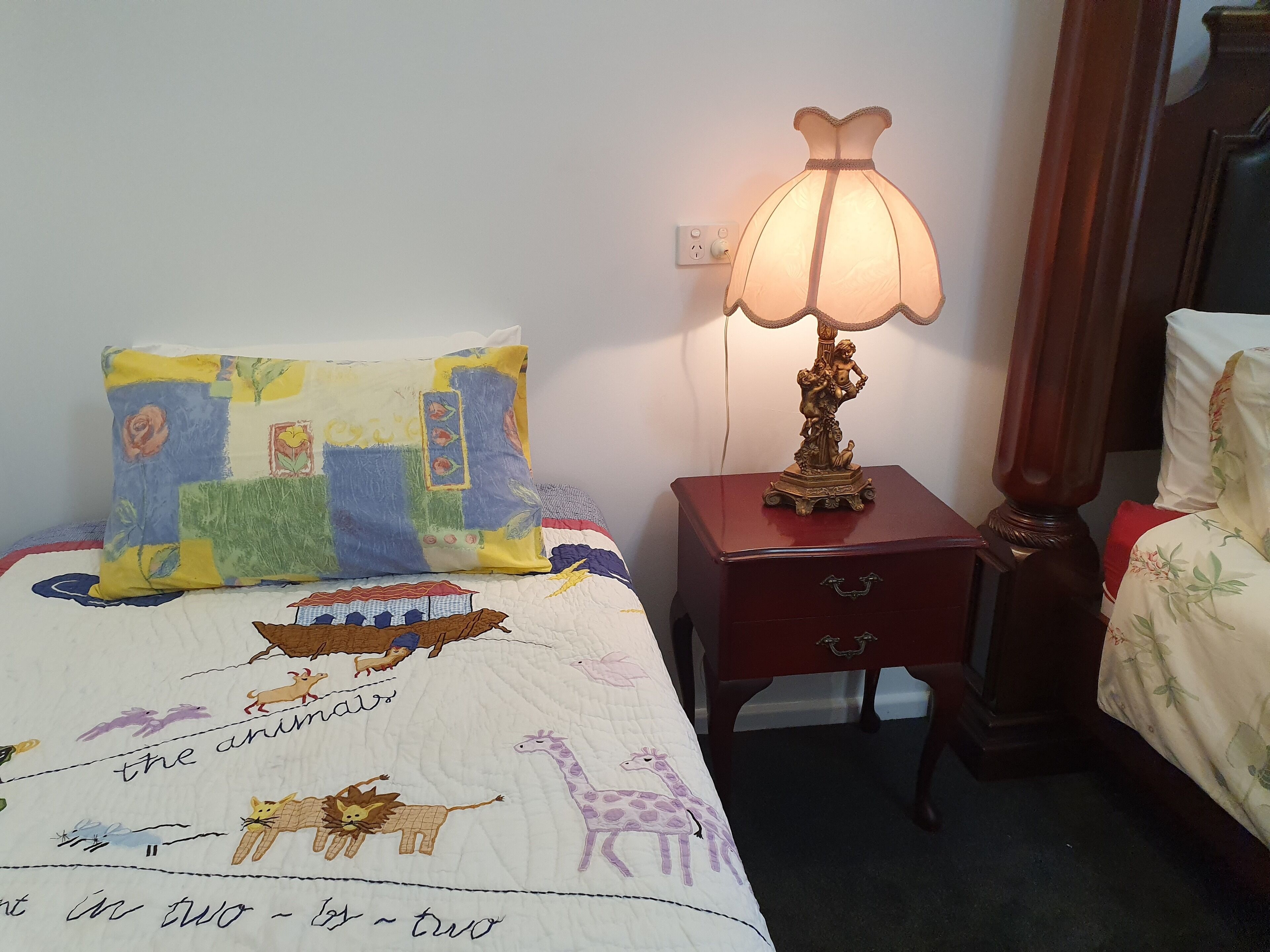 A bedroom with a bed featuring a colorful quilt with animal illustrations, a bedside lamp, a wooden nightstand, and a mirror.