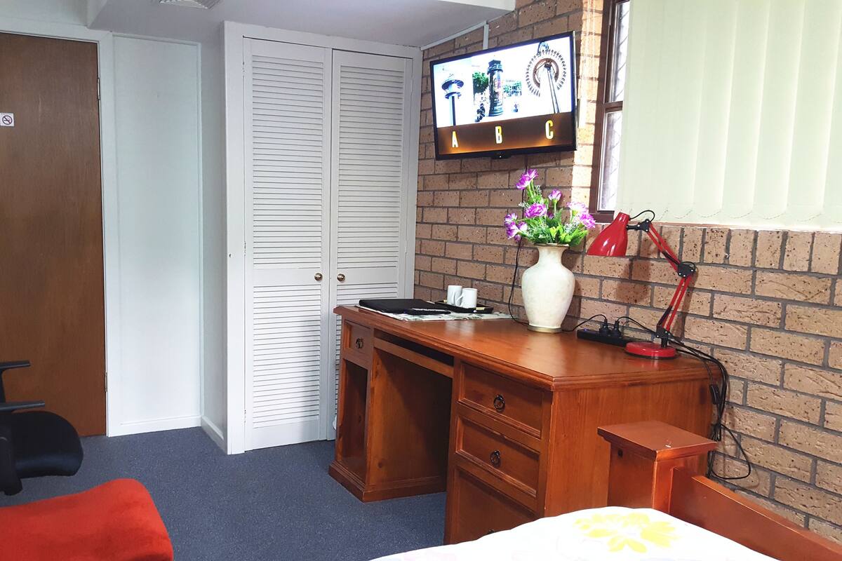 Ein Hotelzimmer mit einem hölzernen Schreibtisch, einem roten Sessel, einem Bett mit floralem Überwurf, einer Backsteinwand, einem über dem Schreibtisch montierten Fernseher und einer Vase mit Blumen auf dem Schreibtisch.