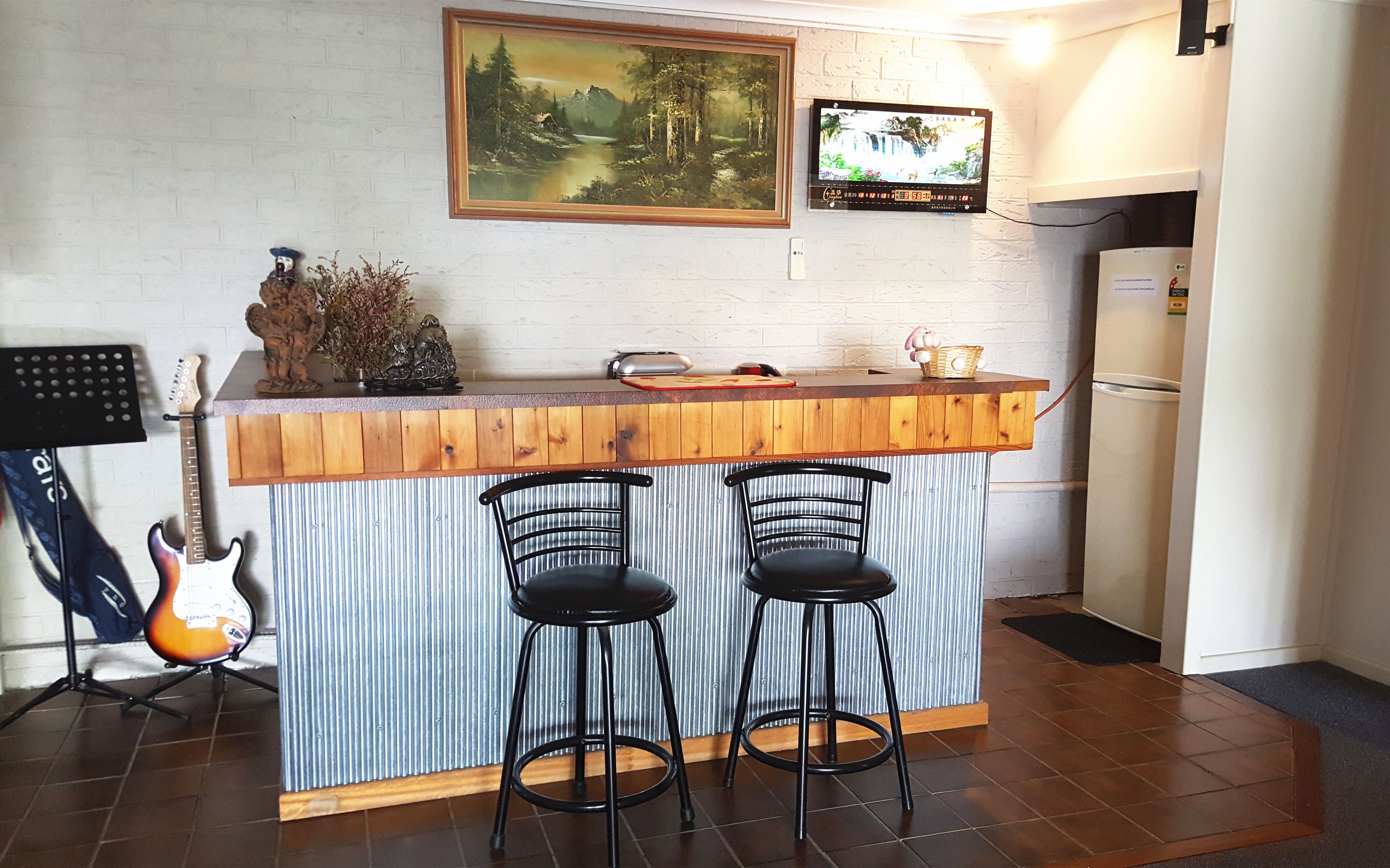 Une cuisine avec un comptoir-bar en bois, deux tabourets de bar noirs, une guitare, un tableau et un téléviseur fixé au mur.