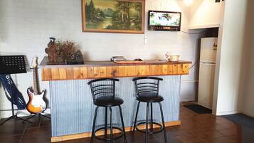 Une cuisine avec un comptoir-bar en bois, deux tabourets de bar noirs, une guitare, un tableau et un téléviseur fixé au mur.