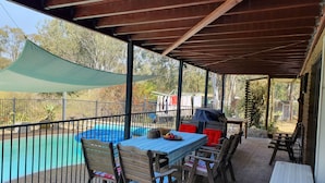 An outdoor patio with a table and chairs, a pool, and a covered area with a view of trees and a house in the background. - Gowrie Agapanthous - Hostel (Gowrie)