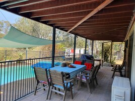 Un patio al aire libre con una mesa y sillas, una piscina y un área cubierta con vista a árboles y una casa al fondo.