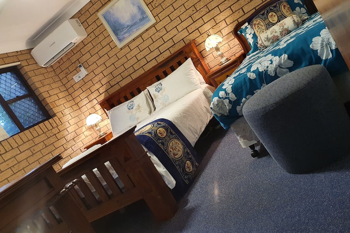 Ein Zimmer mit Backsteinwand, einem Bett mit floralem Bettzeug, einer Sitzbank und einem kleinen Tisch mit Lampe.