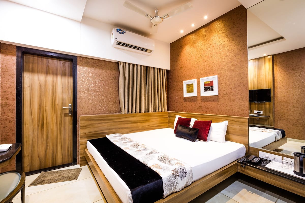 A hotel room with a wooden bed, a desk, a chair, an air conditioning unit, a mirror, and a wall-mounted TV.