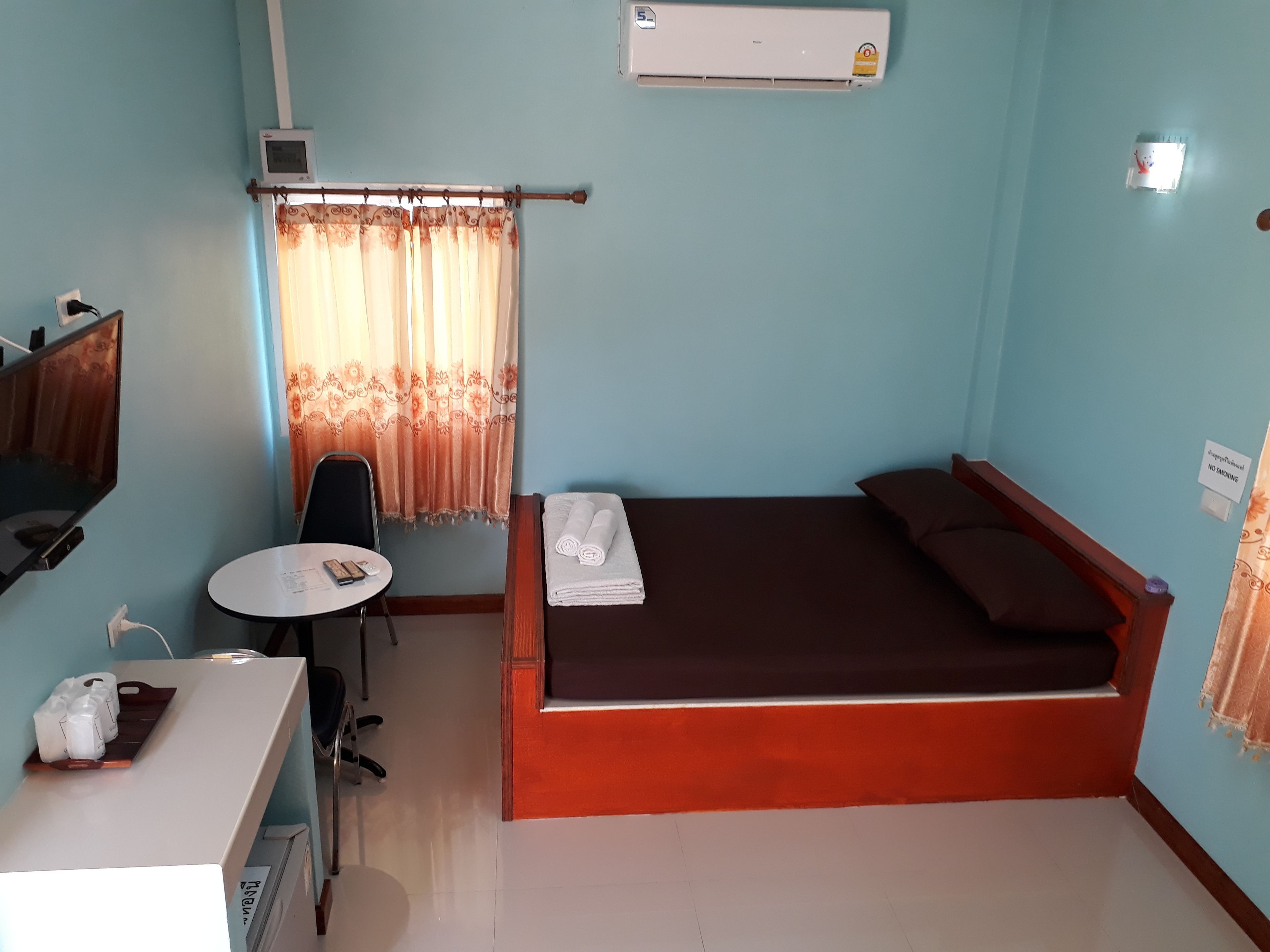 A small, well-lit room with a bed, a desk with a chair, a TV, and an air conditioner.