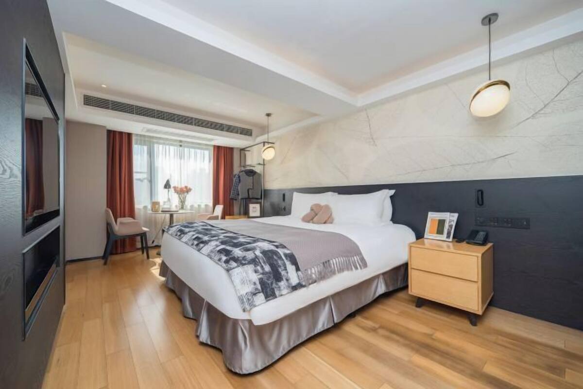 A modern hotel room with a large bed, a bedside table, a TV, and a desk with a chair.