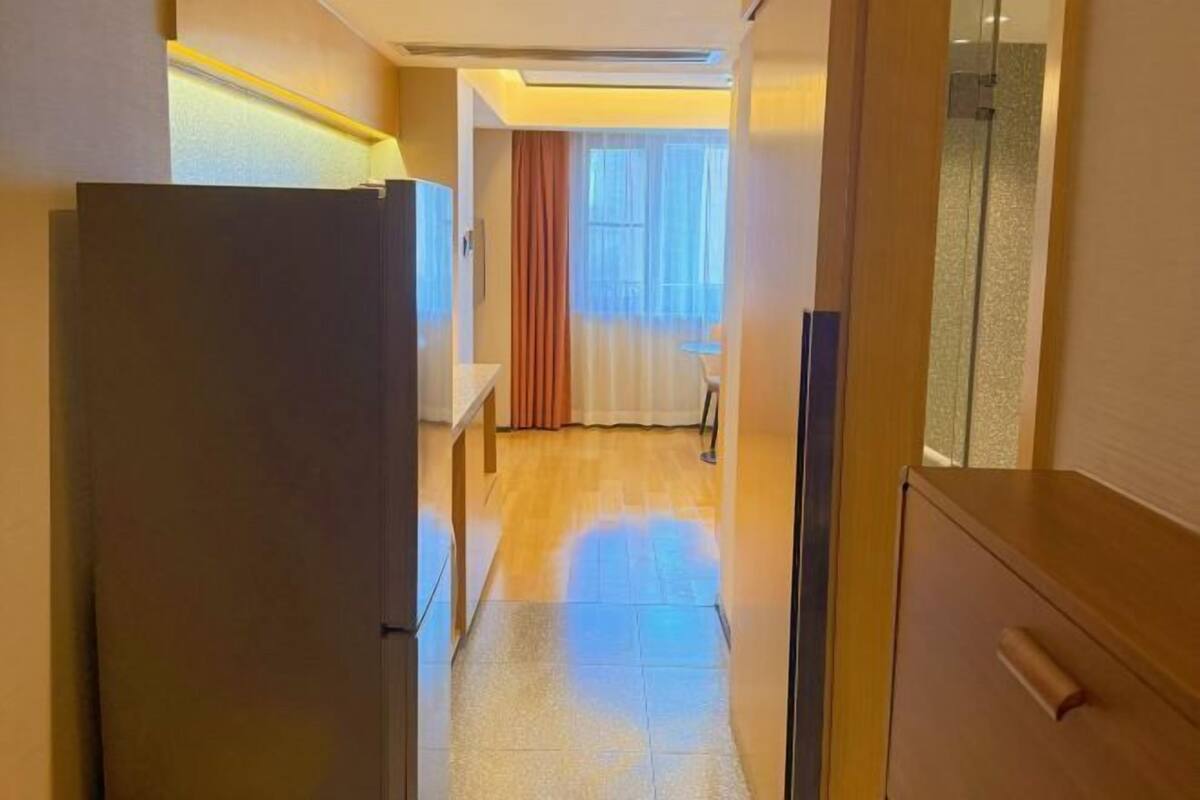 A narrow hallway with a refrigerator on the left, a wooden cabinet on the right, and a window with curtains in the background.