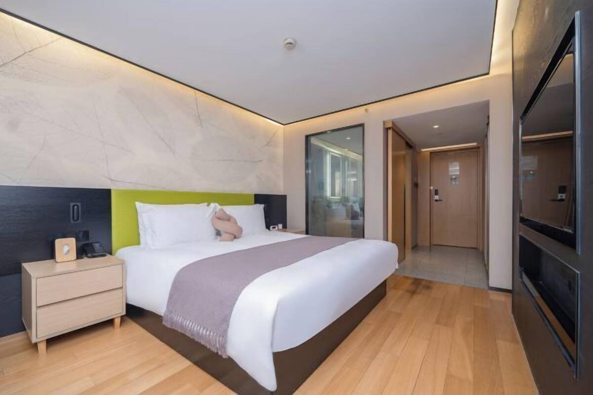 A modern hotel room with a large bed, bedside tables, a TV, and a glass door leading to a bathroom.