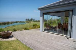 A modern house with a deck overlooking a lake, featuring a glass door leading to an outdoor seating area. - 12 Dunnage (Knysna)
