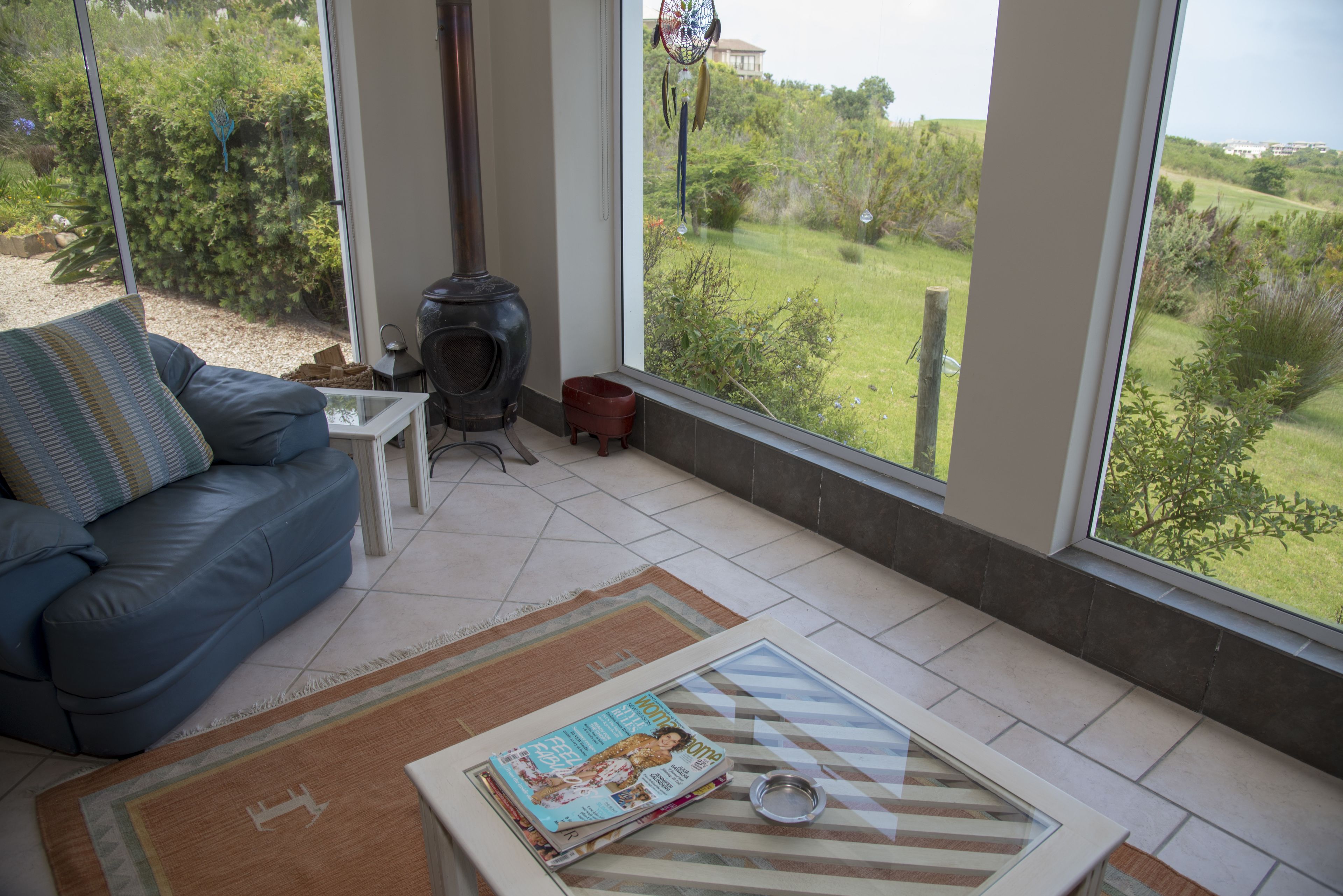 A modern living room with a sofa, a coffee table with a magazine, a small fireplace, and a large window offering a view of a garden.