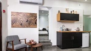 A modern living room with a grey armchair, a small round table, and a zebra-patterned rug. A kitchen with black cabinets and a white countertop is visible in the background. - Kinneret Lodging (Tiberias)